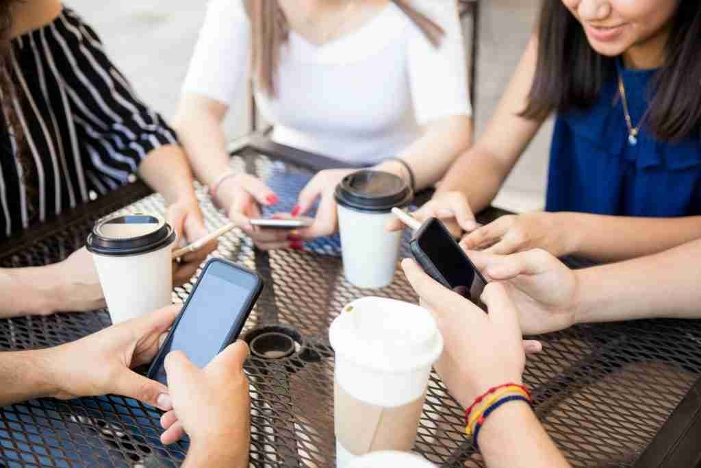 Group of people around a metal table looking at their smartphones with takeaway coffee cups nearby.