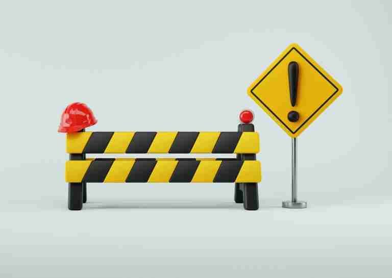 Yellow and black striped construction barrier with a red hard hat on top and a nearby yellow exclamation warning sign.
